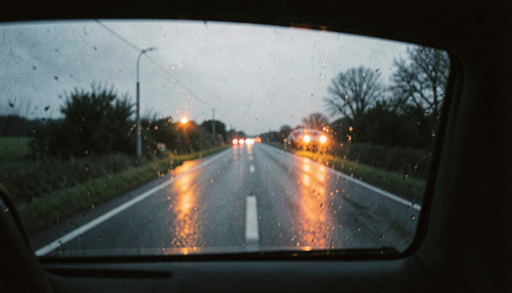 Blick von innen durch eine leicht beschlagene Autoscheibe auf eine nasse Landstraße in der Dämmerung, Scheinwerferreflexionen, gedämpfte Blau- und Grautöne, analoge Filmfotografie.