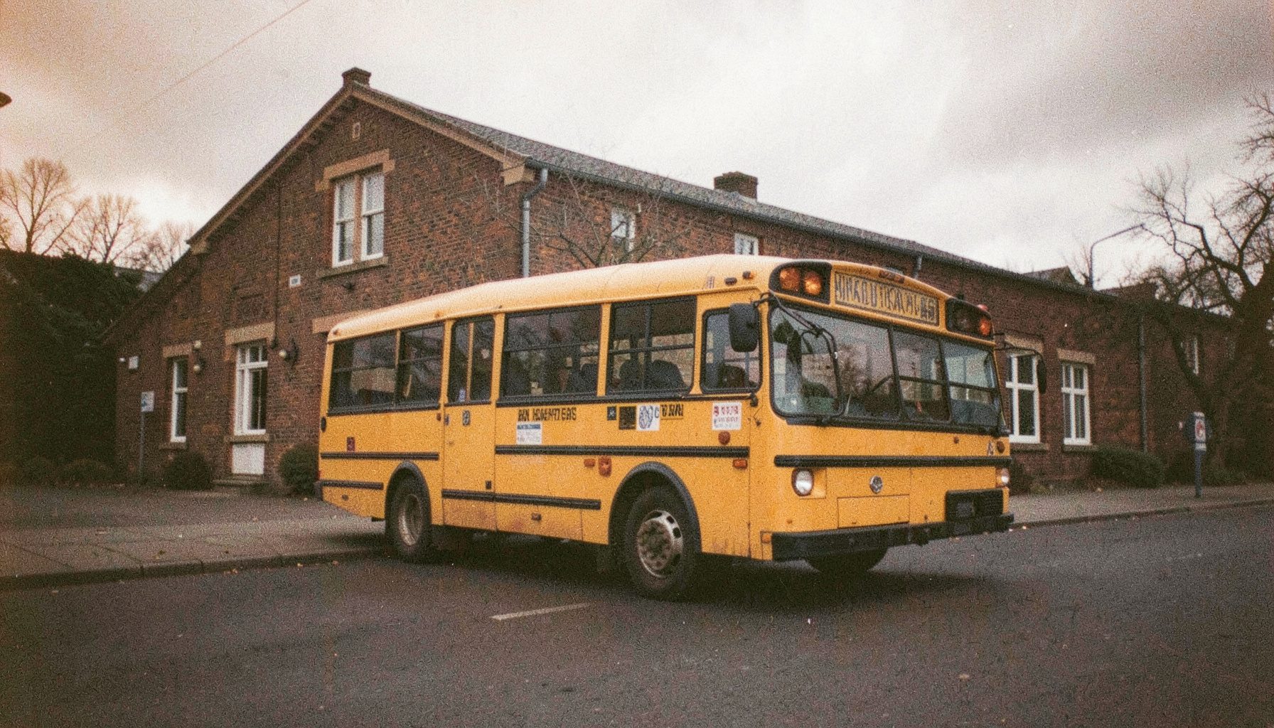Ein gelber Schulbus, geparkt am Straßenrand vor einer Backstein-Schule an einem bewölkten Tag, gedämpftes Licht, analoge Filmfotografie mit sichtbarem Korn.