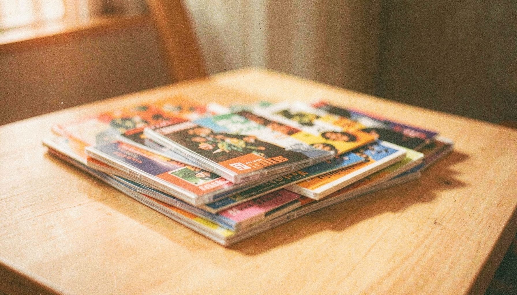 Weichgezeichnetes analoges Filmfoto mit sichtbarem Korn: ein Stapel DVDs und bunter Broschüren auf einem hellen Holztisch, warmes Seitenlicht, ruhige und einladende Stimmung