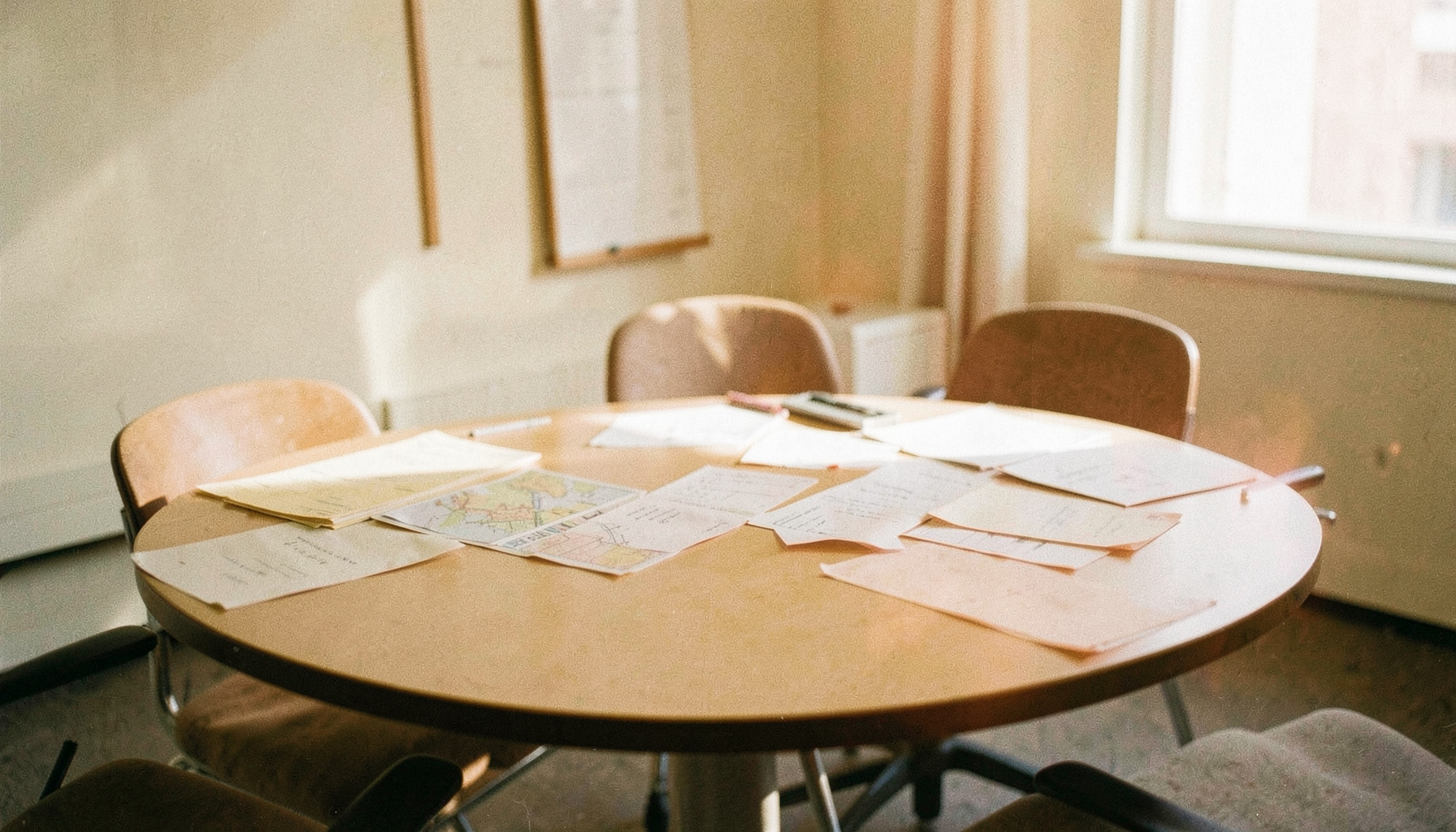 Eine analoge Filmfotografie mit sichtbarem Korn: ein runder Tisch in einem hellen Besprechungsraum, darauf ausgebreitete Karten und Unterlagen, warmes Tageslicht von der Seite, überwiegend cremefarbene und holzbraune Töne, ruhige und sachliche Atmosphäre.